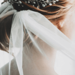 Image shows the head and neck of the white woman with blonde hair from behind. Her hair is in an up style and she is wearing a bridal veil