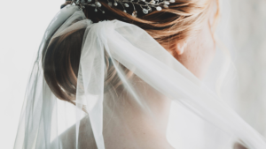 Image shows the head and neck of the white woman with blonde hair from behind. Her hair is in an up style and she is wearing a bridal veil