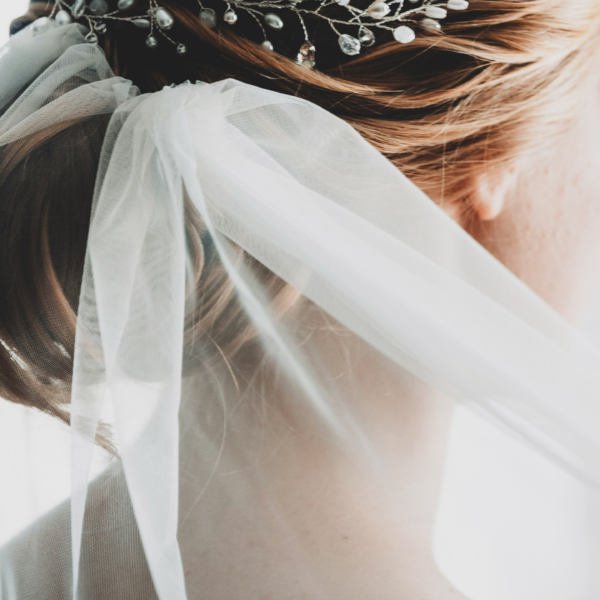 Image shows the head and neck of the white woman with blonde hair from behind. Her hair is in an up style and she is wearing a bridal veil